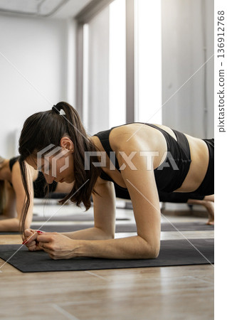 A woman with dark hair in a ponytail performs a forearm plank on a yoga mat in a bright studio, focusing on her core strength A woman with dark hair in a ponytail performs a forearm plank on a yoga mat in a bright studio, focusing on her core strength 136912768