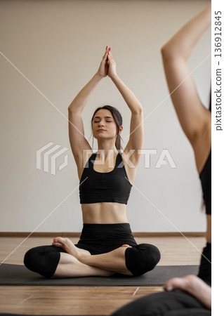A woman with eyes closed practices yoga in a serene studio, holding her hands in prayer above her head while seated in a lotus position 136912845