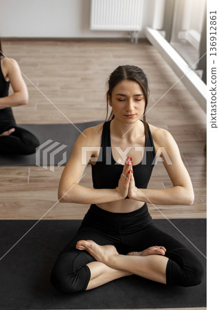 A woman in black activewear sits in a lotus position on a yoga mat, eyes closed with hands pressed together in prayer A woman in black activewear sits in a lotus position on a yoga mat, eyes closed with hands pressed together in prayer 136912861