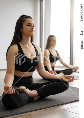 Two women in black activewear practice meditation and mindfulness on yoga mats in a bright studio with large windows 136912867