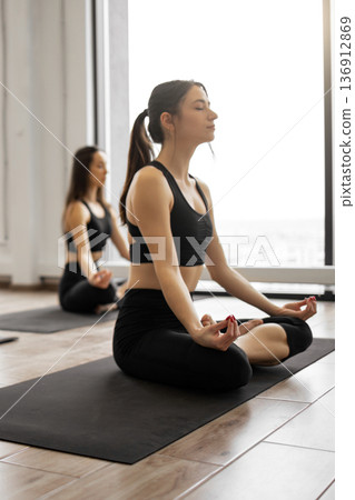 Two women in black activewear practice meditation and yoga poses on mats in a bright studio with large windows Two women in black activewear practice meditation and yoga poses on mats in a bright studio with large windows 136912869