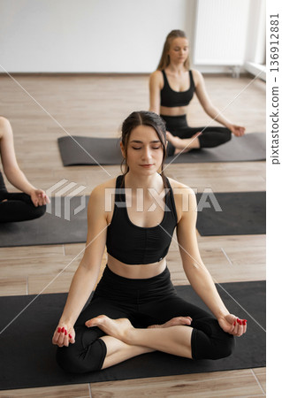 Three women in black activewear practice meditation and mindfulness on yoga mats in a bright studio room Three women in black activewear practice meditation and mindfulness on yoga mats in a bright studio room 136912881