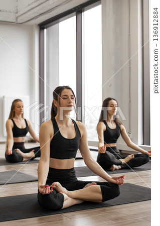 Three women in black activewear practice meditation and mindfulness on yoga mats in a bright studio with large windows 136912884