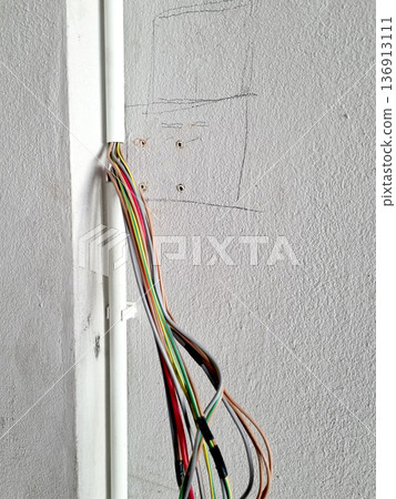 Bundle of colorful electrical wires protruding from a white plastic conduit on a textured concrete wall during home renovation. Includes pencil marks and drill holes for a power socket installation. 136913111