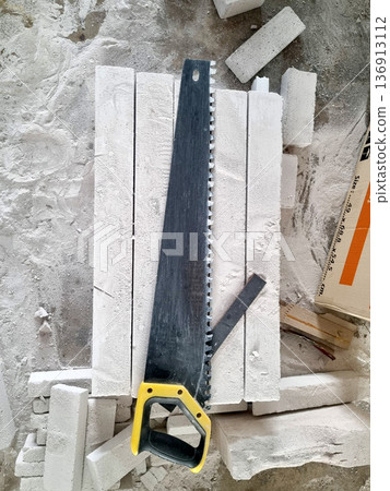 Hand saw with yellow handle resting on stacked aerated concrete blocks at a construction site. High angle view of professional masonry tools and building materials in a dusty environment. 136913112
