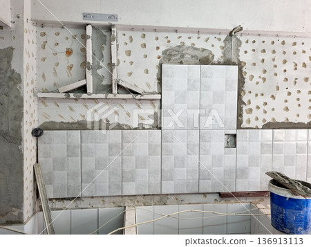 Kitchen wall renovation with grey ceramic tiles being installed over concrete. Professional home improvement process showing construction details, adhesive marks and tile placement on site. 136913113