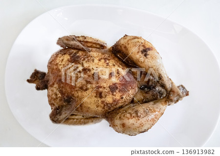 Whole roasted chicken on a white plate, prepared for a meal. Delicious cooked poultry dish for dinner or holiday food concept. Whole roasted chicken on a white plate, prepared for a meal. Delicious cooked poultry dish for dinner or holiday food concept. 136913982