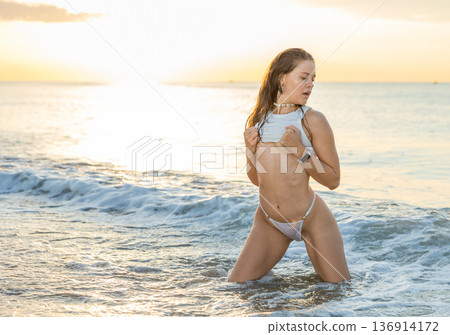 Young woman in thong and top posing while kneeling on beach Young woman in thong and top posing while kneeling on beach 136914172