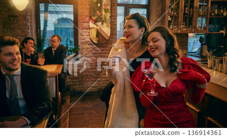 Glamorous women in evening dresses enjoying cocktails at retro bar. Glamorous women in evening dresses enjoying cocktails at retro bar. 136914361