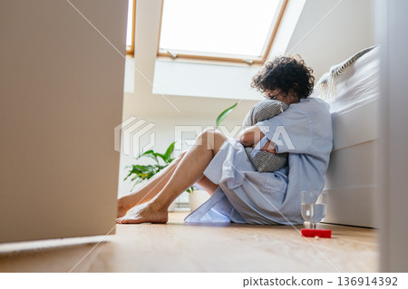 Despaired woman with curly hair and glasses sitting barefoot on bedroom floor near bed, tightly hugging pillow, glass of water and antidepressants, mental health crisis at home 136914392
