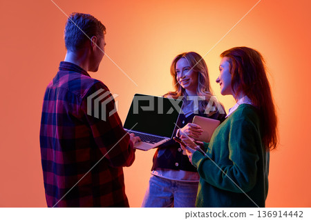 Three young professionals discussing project using laptop and tablet on gradient background. Three young professionals discussing project using laptop and tablet on gradient background. 136914442