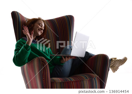 Smiling woman waving during laptop video call in striped armchair. 136914454