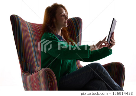 Young woman using tablet while sitting in striped armchair isolated on white. Young woman using tablet while sitting in striped armchair isolated on white. 136914458