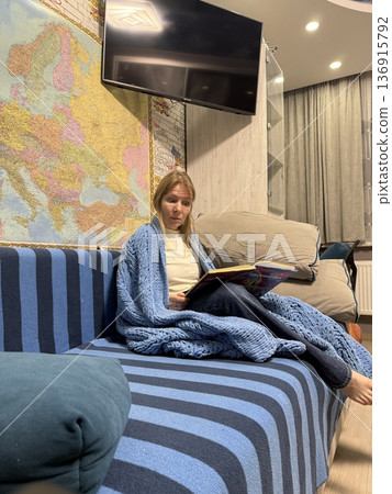 A woman wrapped in a blue knitted blanket sits on a striped sofa and reads a book, cozy lifestyle, reading hobby, peaceful moment. A woman wrapped in a blue knitted blanket sits on a striped sofa and reads a book, cozy lifestyle, reading hobby, peaceful moment. 136915792