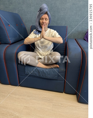 A woman with a blue towel on her head sits on a blue sofa with hands in a prayer pose, morning wellness, meditation practice, home spa. 136915795