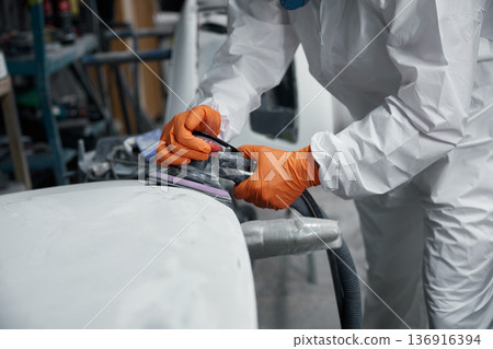 Auto body technician in a protective suit meticulously sands a car hood at a repair shop 136916394