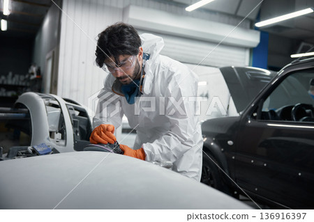 Auto body technician in a protective suit meticulously sands a car hood at a repair shop 136916397