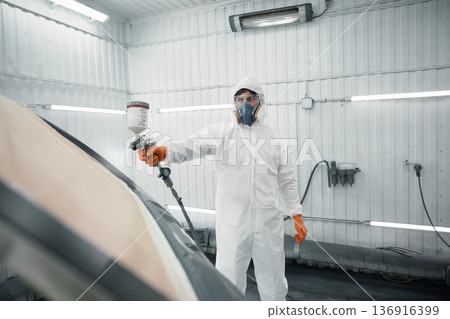 An auto painter in a protective suit is skillfully spraying a car in a paint booth An auto painter in a protective suit is skillfully spraying a car in a paint booth 136916399