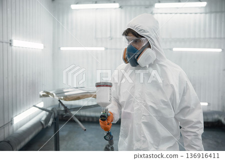 A worker dressed in a protective suit is spray-painting a car panel inside a modern paint booth A worker dressed in a protective suit is spray-painting a car panel inside a modern paint booth 136916411