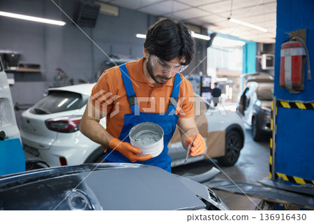 Technician is meticulously prepping a car panel with primer in a garage workshop 136916430