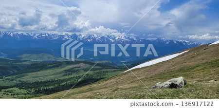Scenic view of a mountain slope with patches of snow and grass, set against a backdrop of distant, snow-capped mountains under a cloudy sky. Scenic view of a mountain slope with patches of snow and grass, set against a backdrop of distant, snow-capped mountains under a cloudy sky. 136917122