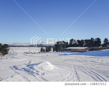 Winter landscape with expansive snow-covered field and mountain backdrop. Rural scenery with evergreen trees and building in cold season. Winter landscape with expansive snow-covered field and mountain backdrop. Rural scenery with evergreen trees and building in cold season. 136917211