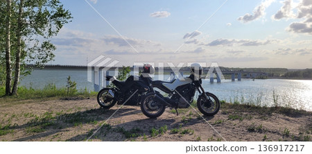 Two motorcycles with helmets parked on sandy ground near a river and a bridge. Travel and adventure concept for motorcycling lifestyle. Two motorcycles with helmets parked on sandy ground near a river and a bridge. Travel and adventure concept for motorcycling lifestyle. 136917217