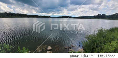 Expansive lake under a cloudy sky, surrounded by dense green forest foliage, showcasing natural beauty and tranquil water surface. 136917222