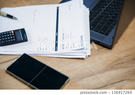 Stack of invoice documents on a wooden desk with a calculator, a pen, a phone, and a laptop. Business accounting concept for financial management. 136917269