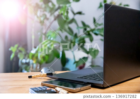 Laptop, phone, pen, and mouse on a wooden desk with a green plant in the background, representing remote work space and modern technology. 136917282