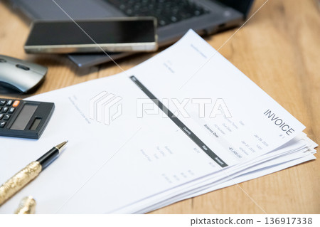 Invoice document stacked on wood table with calculator, pen, smartphone, and laptop. Financial management, business accounting. 136917338