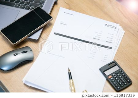 Invoice document on a wooden desk with a calculator, pen, smartphone, and laptop. Financial management and payment processing concept. 136917339