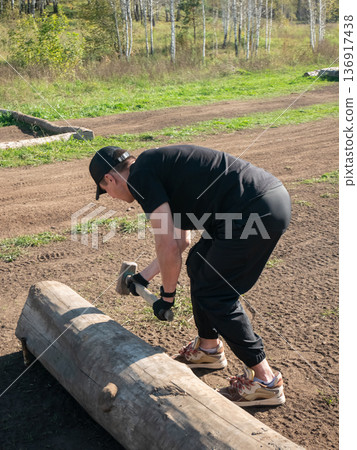 Young man drives in a log with a mallet outdoors. Fitness or outdoor physical workout concept. For health, training, lifestyle. Young man drives in a log with a mallet outdoors. Fitness or outdoor physical workout concept. For health, training, lifestyle. 136917438