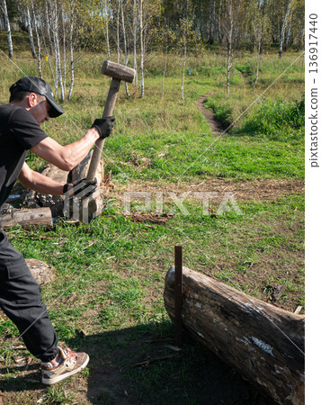 Man splitting wood with a large hammer outdoors. Work and chores concept in a natural environment during daytime. Manual labor. Man splitting wood with a large hammer outdoors. Work and chores concept in a natural environment during daytime. Manual labor. 136917440