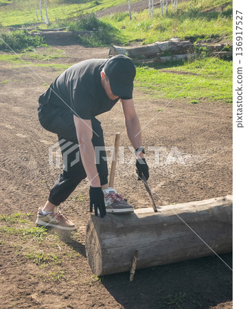 Man aligning wooden log on dirty ground with stick, wearing black cap and gloves for construction work or landscaping project Man aligning wooden log on dirty ground with stick, wearing black cap and gloves for construction work or landscaping project 136917527