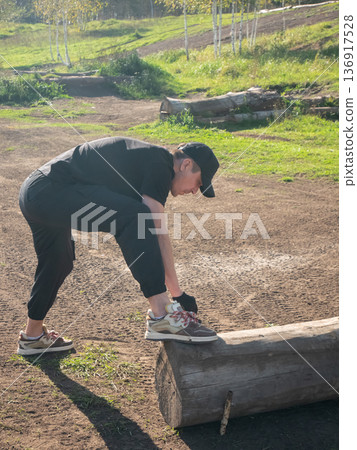Man tying shoelace outdoors with his foot on a log. Preparation for training or outdoor activity. Active lifestyle concept. Man tying shoelace outdoors with his foot on a log. Preparation for training or outdoor activity. Active lifestyle concept. 136917528
