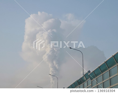 Thick smoke plume rises into the sky from an industrial source behind a highway sound barrier, symbolizing environmental pollution. Thick smoke plume rises into the sky from an industrial source behind a highway sound barrier, symbolizing environmental pollution. 136918304