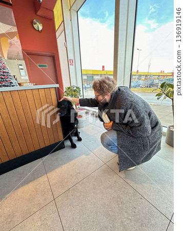 Man touching black dog inside cafe near window. Human animal bond, companionship care, kindness gesture, everyday urban life concept 136919146