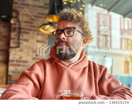 Bearded man in glasses drinking tea at cafe window. Urban lifestyle, contemplation mood, social space, modern city living concept 136919154
