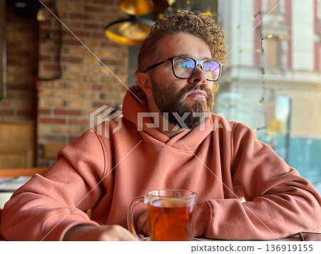 Bearded man in glasses drinking tea at cafe window. Urban lifestyle, contemplation mood, social space, modern city living concept Bearded man in glasses drinking tea at cafe window. Urban lifestyle, contemplation mood, social space, modern city living concept 136919155