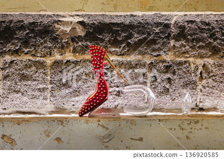 A red high heel shoe is on a ledge next to a wine glass. A red high heel shoe is on a ledge next to a wine glass. 136920585