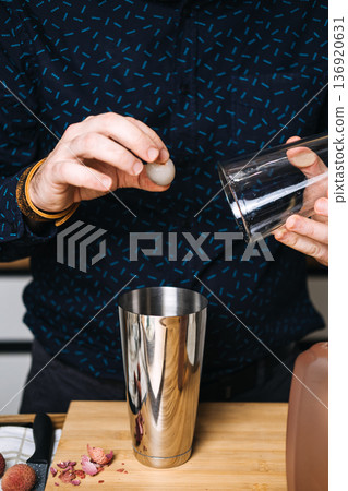 Bartender drops peeled lychee into stainless steel cocktail shaker next to fruit skins and glass. Craft cocktail movement, professional bartender, mixology techniques, lychee martini, artisan drinks. 136920631