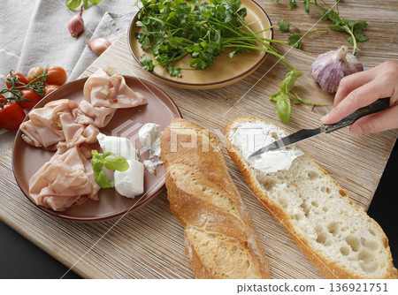 Anonymous person spreads soft goat cheese on bread, preparing basil and mortadella sandwich in kitchen. Italian cuisine, high-quality farm-to-table produce. Anonymous person spreads soft goat cheese on bread, preparing basil and mortadella sandwich in kitchen. Italian cuisine, high-quality farm-to-table produce. 136921751