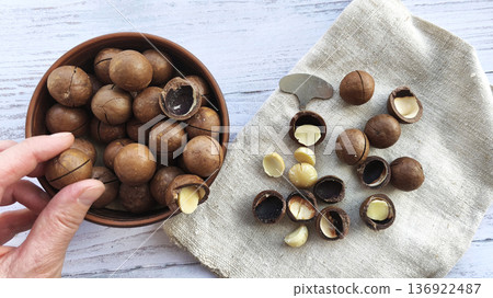Macadamia nuts in a bowl with cracked shells and kernels on linen cloth. Hand picking nut, nutcracker tool nearby. Natural organic snack preparation, rustic flat lay food background. Macadamia nuts in a bowl with cracked shells and kernels on linen cloth. Hand picking nut, nutcracker tool nearby. Natural organic snack preparation, rustic flat lay food background. 136922487
