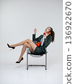 An elegant white woman in a green business suit talks on a red telephone while sitting on a chair against the backdrop of an isolated space. Office. Curly hair, brown-haired 136922670