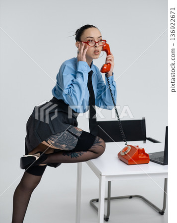 Young calm white woman in office attire working talking on vintage red phone, isolated on white background. In a blue shirt with a tie. Business occupation. Young calm white woman in office attire working talking on vintage red phone, isolated on white background. In a blue shirt with a tie. Business occupation. 136922671