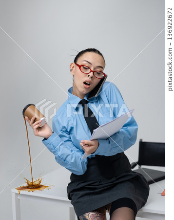 Young white woman in office is talking on the phone holding a cup of coffee and a folder with documents sitting on the edge of a table in business attire Blue shirt glasses Isolated space Pours coffee Young white woman in office is talking on the phone holding a cup of coffee and a folder with documents sitting on the edge of a table in business attire Blue shirt glasses Isolated space Pours coffee 136922672
