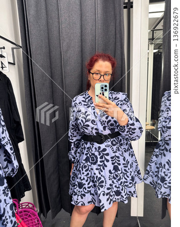 Woman trying patterned dress in fitting room mirror. Fashion choice, retail shopping, style evaluation and modern consumer lifestyle Woman trying patterned dress in fitting room mirror. Fashion choice, retail shopping, style evaluation and modern consumer lifestyle 136922679