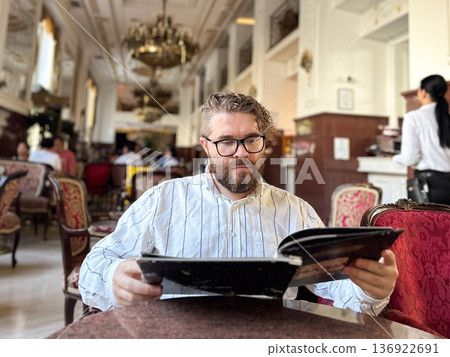 Millennial man in a classic interior reads a menu in a historic restaurant with ornate decor and a grand chandelier. Atmosphere of elegance and timeless charm Millennial man in a classic interior reads a menu in a historic restaurant with ornate decor and a grand chandelier. Atmosphere of elegance and timeless charm 136922691