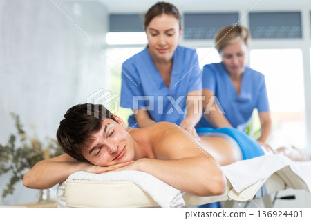 Young man relaxing while two masseuses doing four hands back massage 136924401